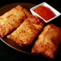 A small round plate holds several halifax style meat paste egg rolls, next to a small bowl of deep orange plum sauce.