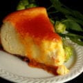 A slice of hop cheesecake with citrus glaze. The cheesecake is very pale yellow, topped with a bright orange glaze. It sits on a white plate, with a piece of fresh hop bine next to it.