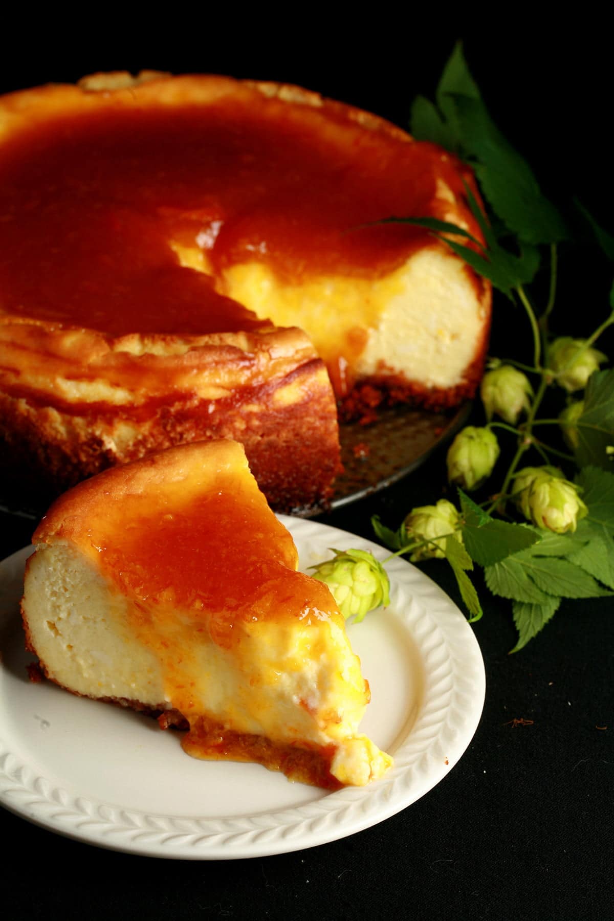 A slice of hopped cheesecake with citrus glaze. The cheesecake is very pale yellow, topped with a bright orange glaze. It sits on a white plate, with a piece of fresh hop bine next to it, with the remainder of the cheesecake in the background.