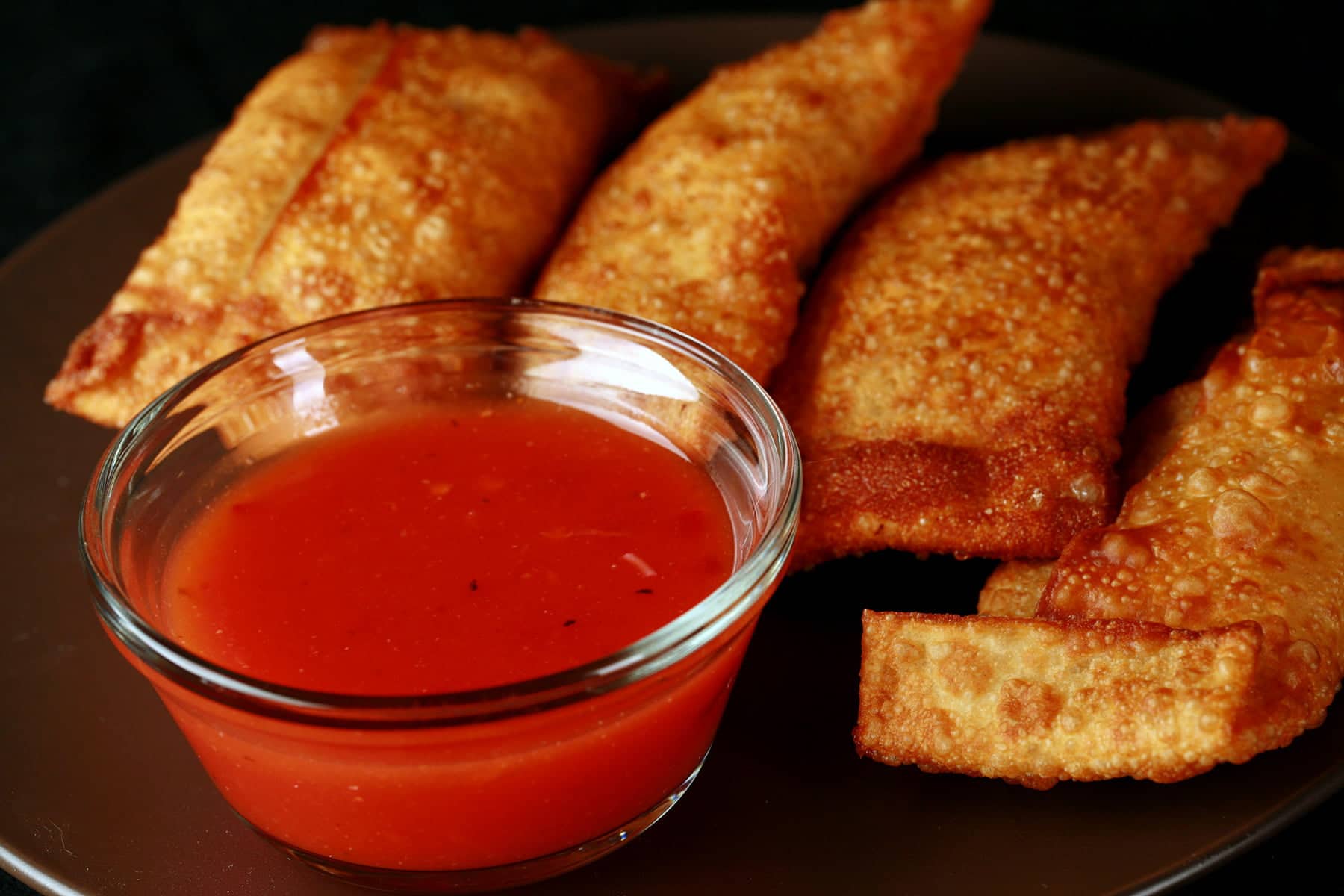A small glass bowl of deeply orange plum sauce is in the foreground, with several egg rolls behind it.