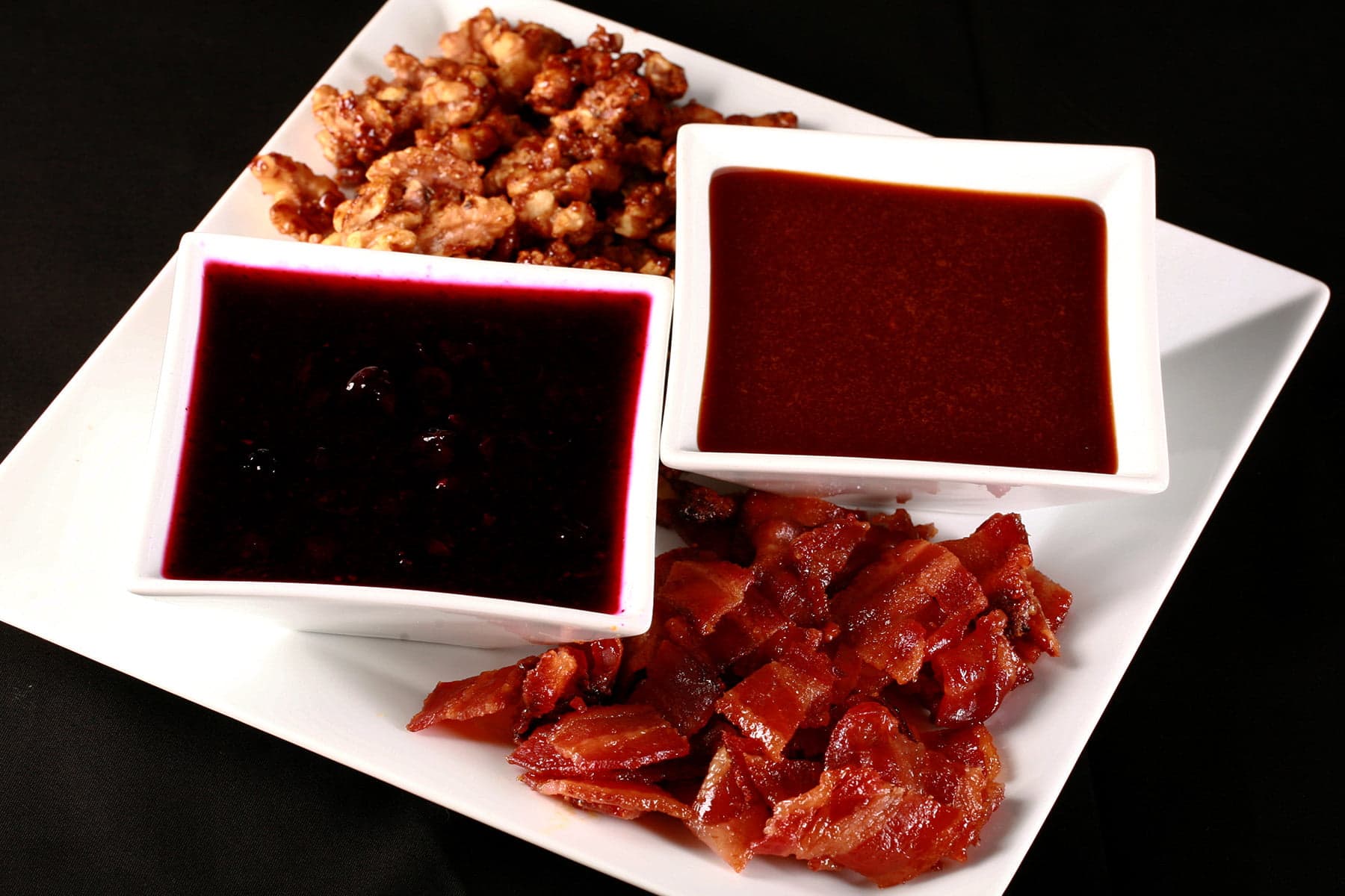 A large square white plate with ice cream toppings arranged on it.  Two square bowls hold sauces - blueberry and salted caramel - while candied bacon is in one corner, and candied nuts are piled in another corner.