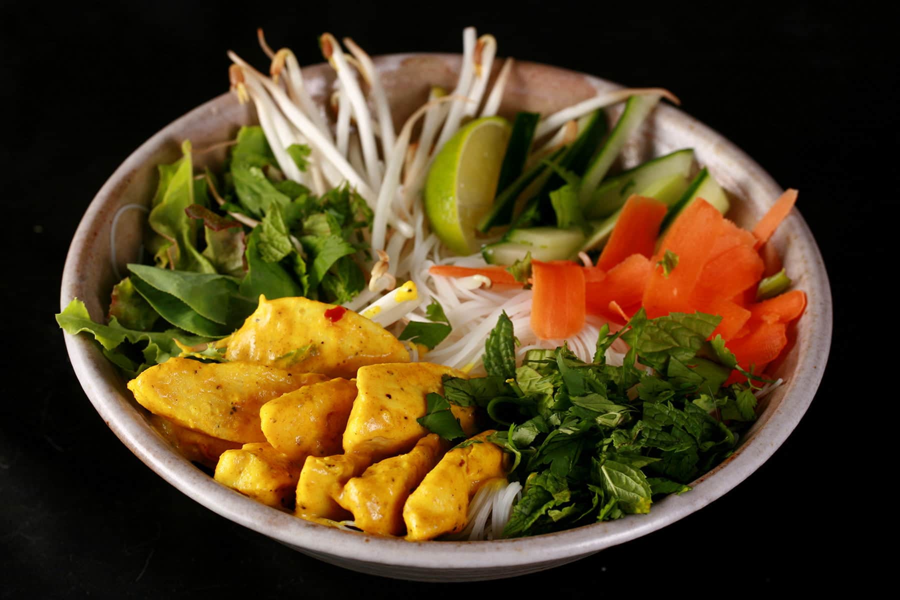A close up view of a bowl of curried chicken Vietnamese noodle salad. Thin vermicelli noodles are visible under an arrangement of curred chicken pieces, cilantro and mint, carrot ribbons, bean sprouts, and lime wedges.