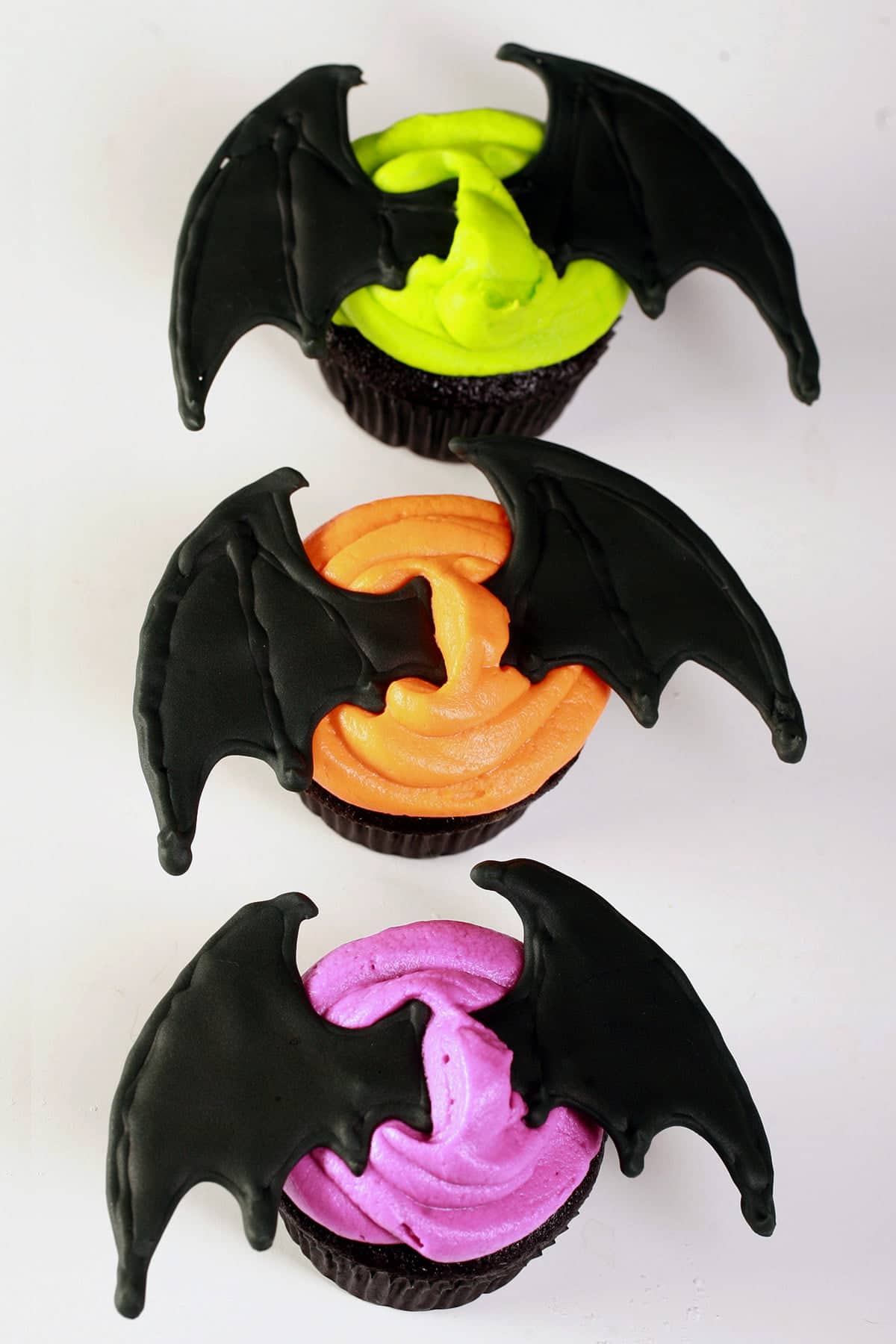 2 Black velvet cupcakes, frosted with bright icing - electric purple, lime green, and orange - and topped with a 3D pair of royal icing bat wings.