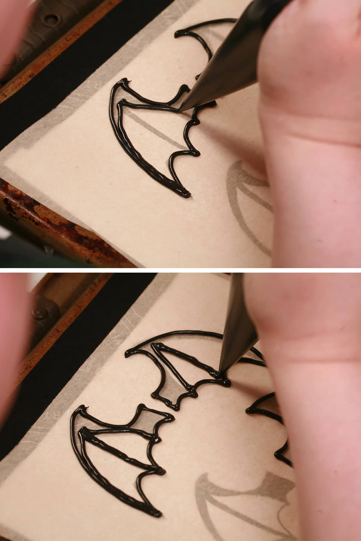 A hand uses a pastry bag of black royal icing to pipe bat wings on a parchment lined baking sheet.