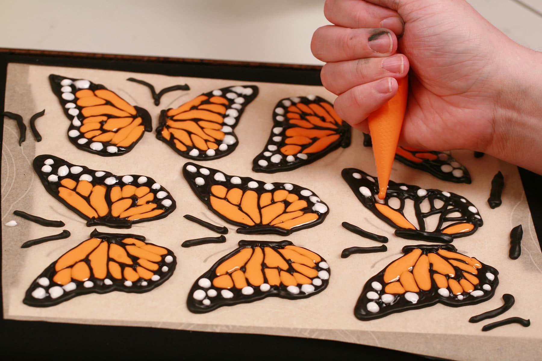 A hand uses a pastry bag full of orange royal icing to fill in the outlines of a monarch butterfly wing.