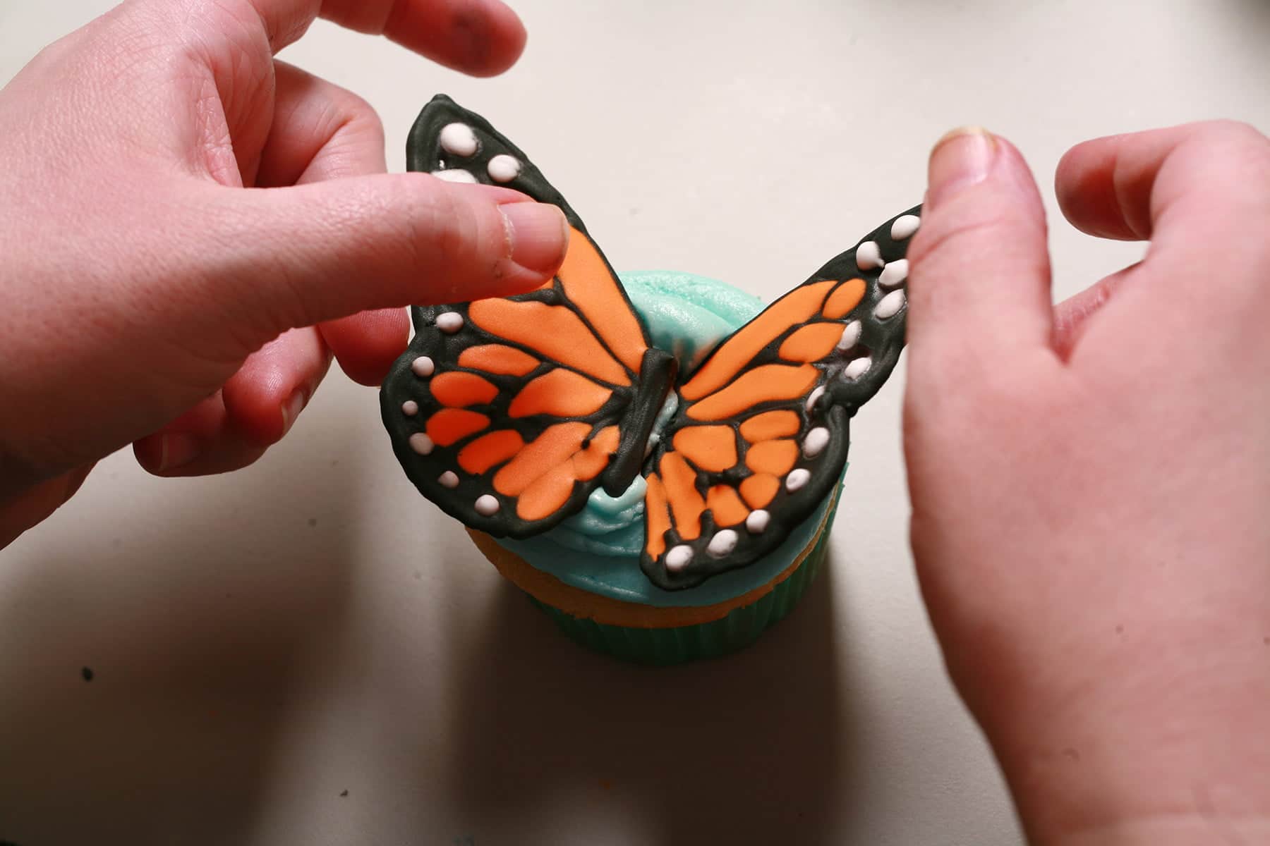 Two hands place a set of monarch butterfly wings on a cupcake iced with blue frosting.