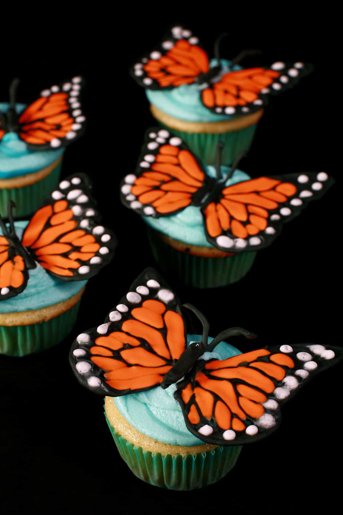 Several 3D Monarch Butterfly Cupcakes, on a black background.  Vanilla cupcakes, frosted with sky blue icing, and topped with Monarch butterfly wings made from royal icing.