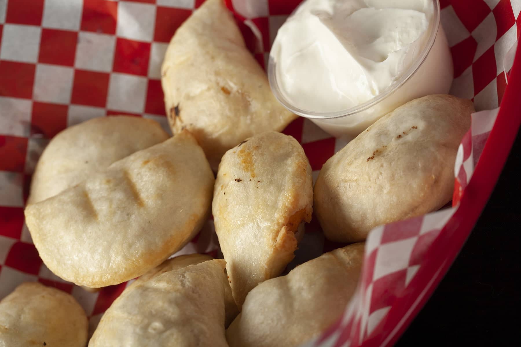 Perfectly fried perogies are mounded in a red basket, accompanied by a small bowl of sour cream.