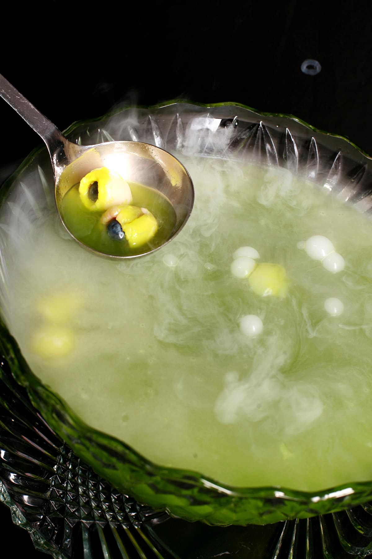 A glass punch bowl with bright green halloween punch, and lychee eyeballs floating in it. Vapour is rising from the punch bowl, from the dry ice used in it.