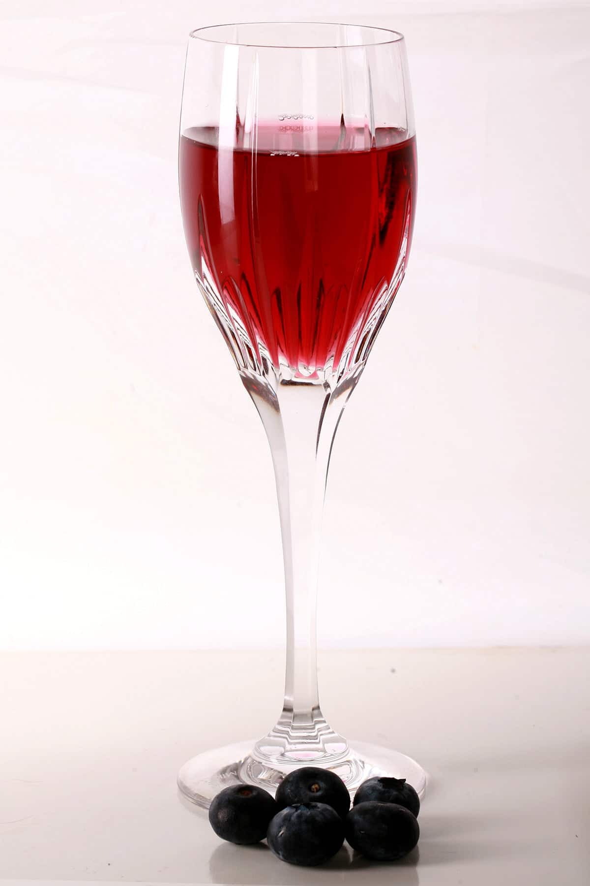 A faceted wine glass full of blueberry mead, with fresh blueberries at the base of the glass.