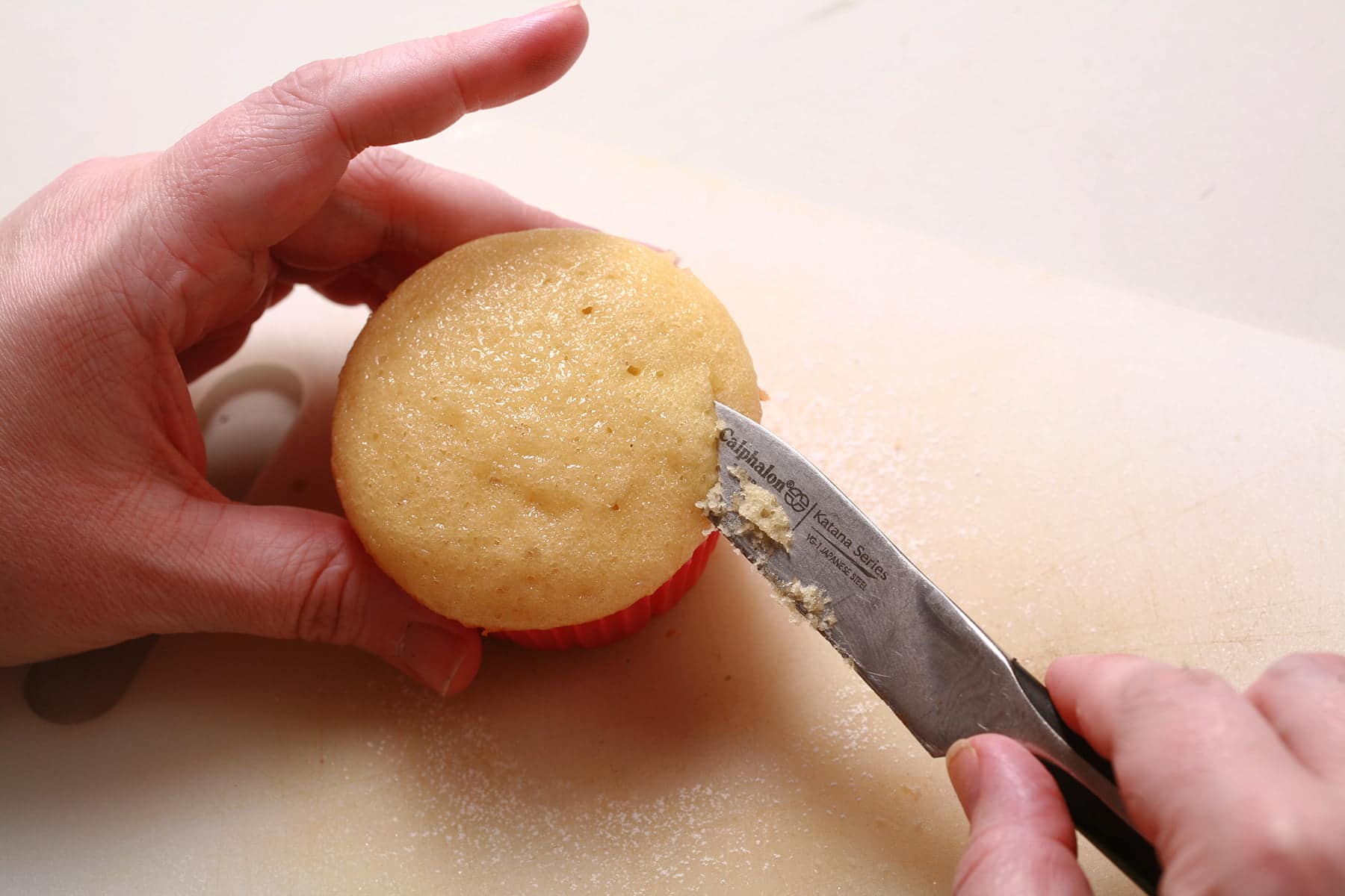 A hand uses a small paring knife to cut the top off a vanilla cupcake