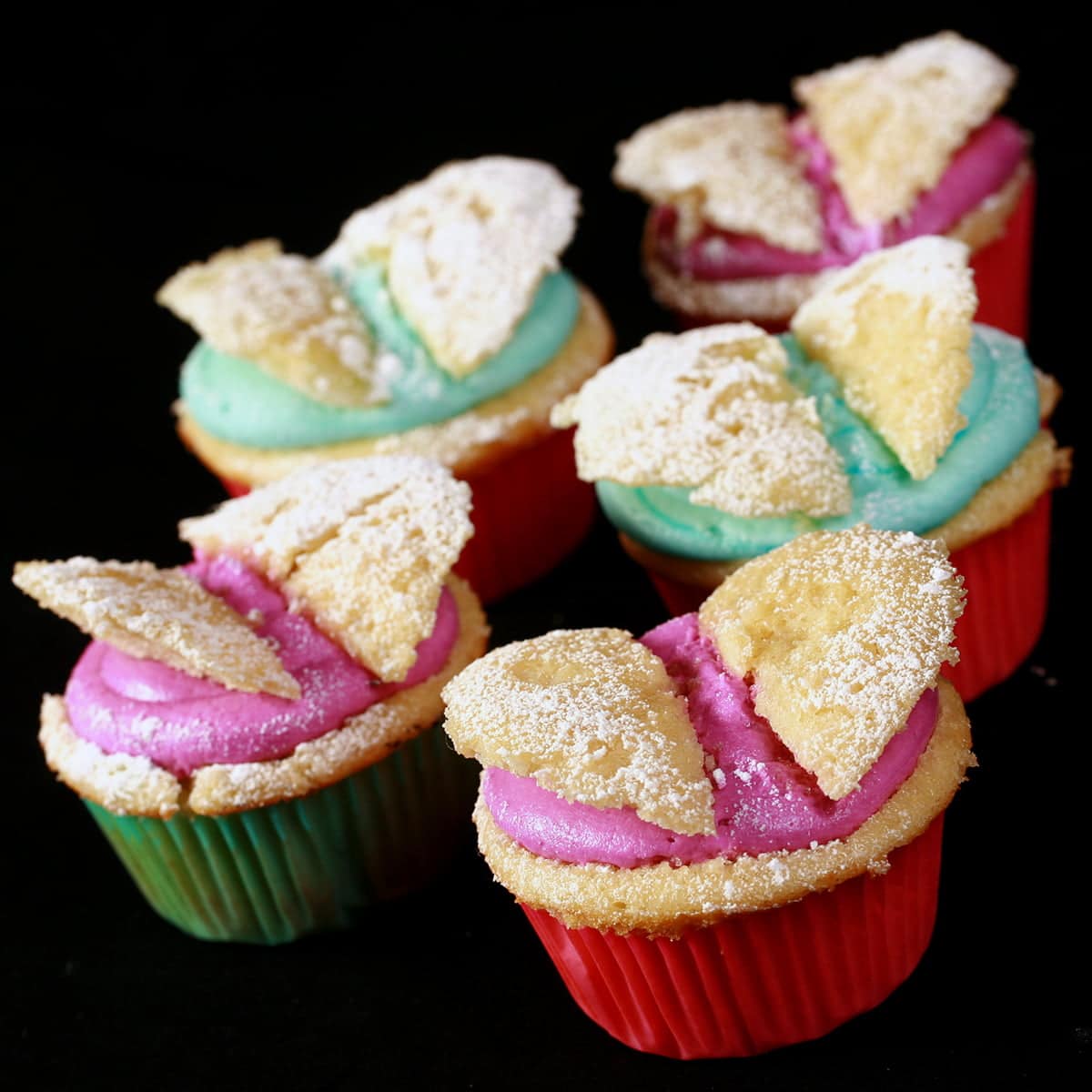 A close up view of several vanilla butterfly cupcakes. The top of each has been cut off, halved, and placed in brightly coloured frosting - teal and magenta - to form wings.
