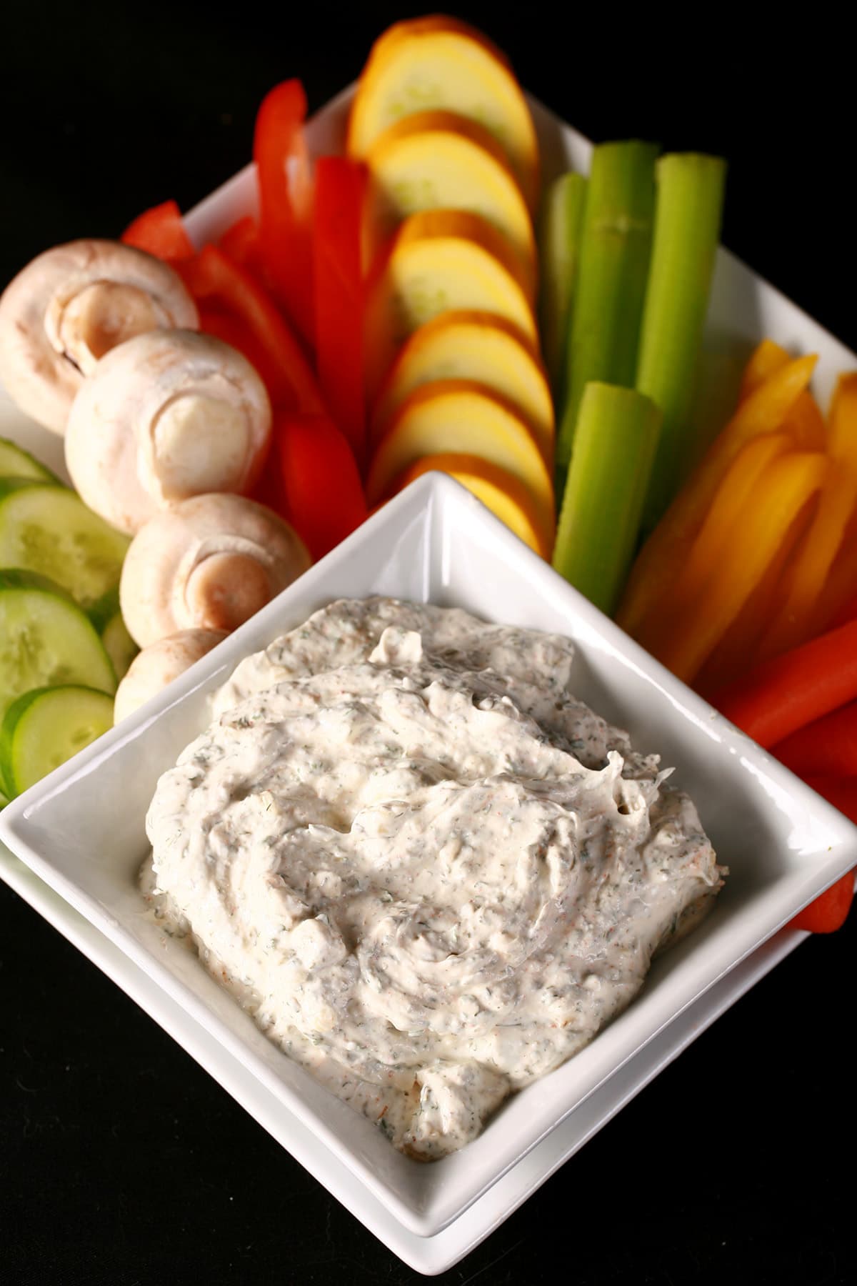 A large white square bowl of easy dill dip, on a larger plate surrounded by a variety of brightly coloured vegetable spears and slices.