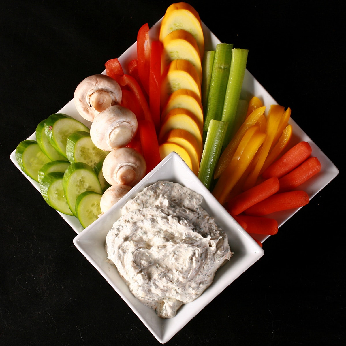 A large white square bowl of easy dill dip, on a larger plate surrounded by a variety of brightly coloured vegetable spears and slices.