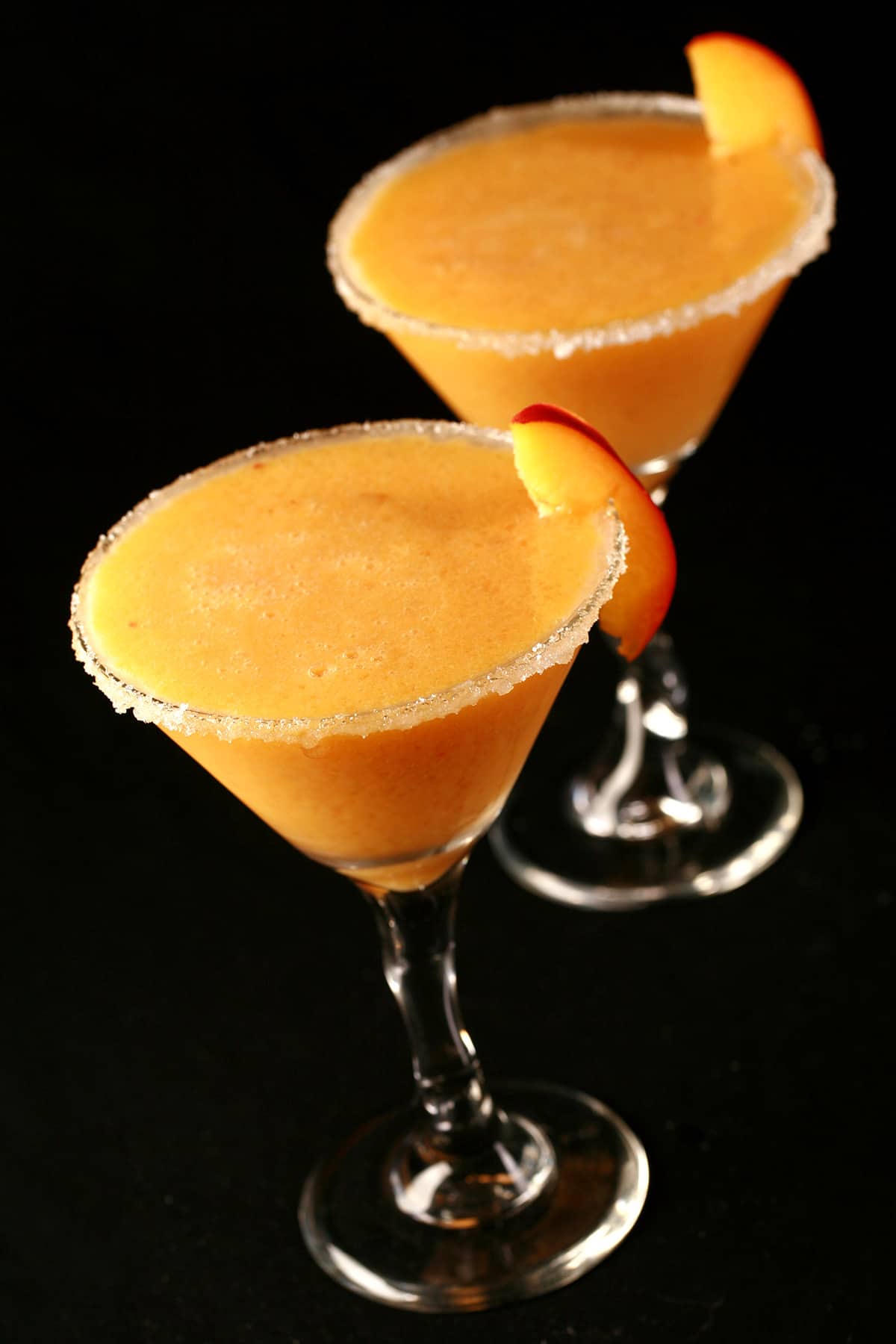 Two Fresh Peach Daiquiris in martini cocktail glasses. The glasses are rimmed with sugar and garnished with a peach slice.