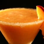 A close up view of a daiquiri made from fresh peaches. It's in a martini cocktail glass rimmed with sugar and garnished with a slice of peach.