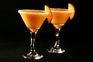 Two Fresh Peach Daiquiris in martini cocktail glasses. The glasses are rimmed with sugar and garnished with a peach slice.