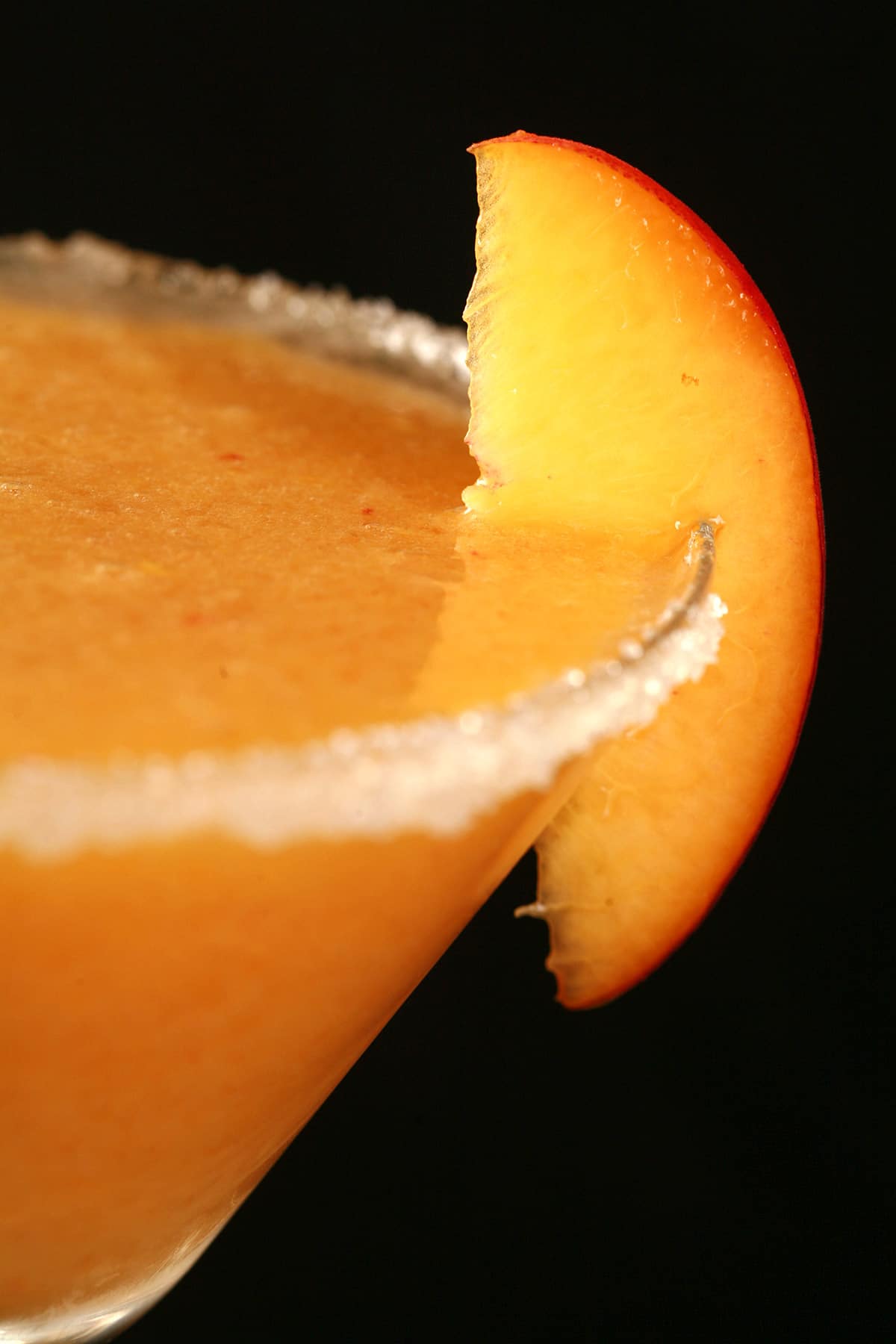 A close up view of a daiquiri made from fresh peaches. It’s in a martini cocktail glass rimmed with sugar and garnished with a slice of peach.