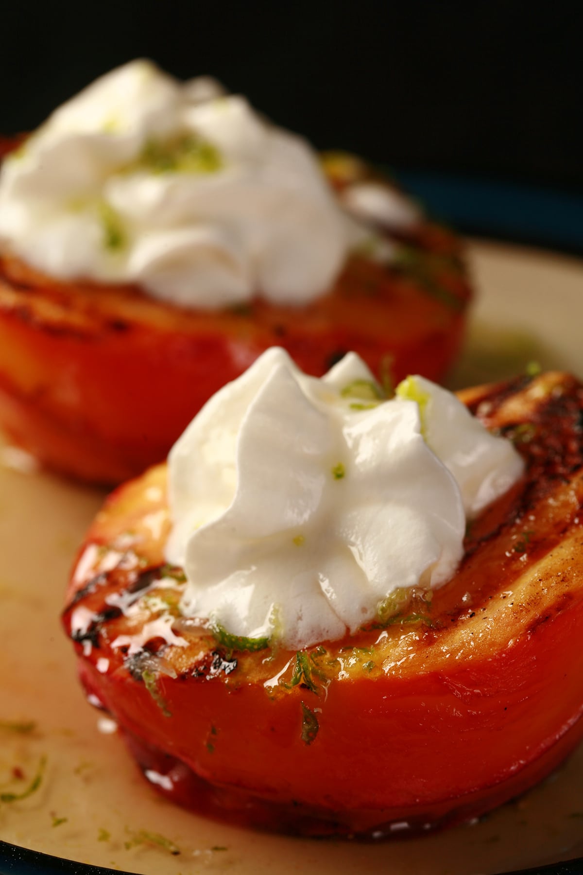 Two grilled peach halves on a plate, drizzled with lime honey. Each has a dollop of whipped cream on it.