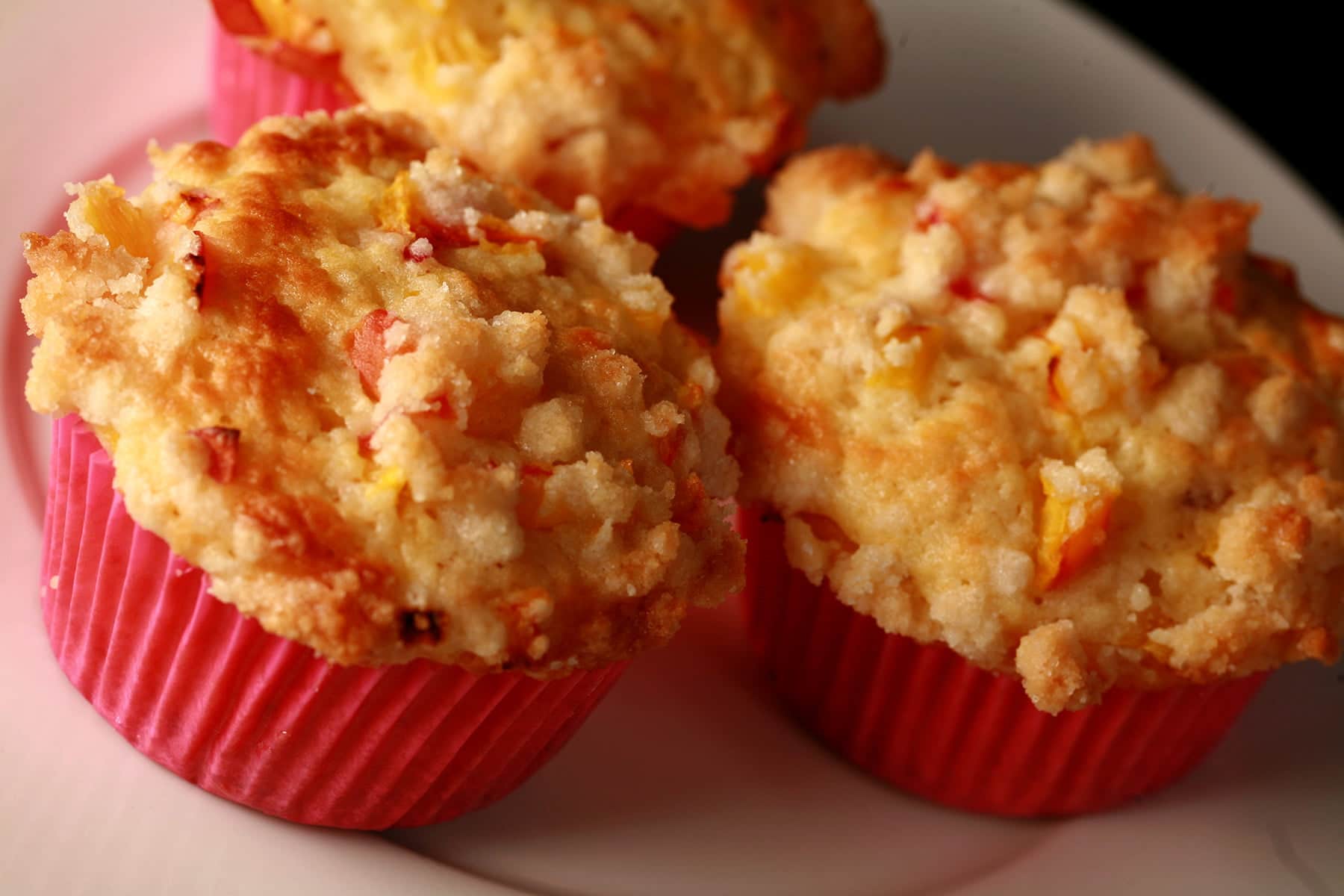 3 Peach cobbler muffins on a white plate. The muffins have small pieces of peach and streusel on top, and each has a pink liner.