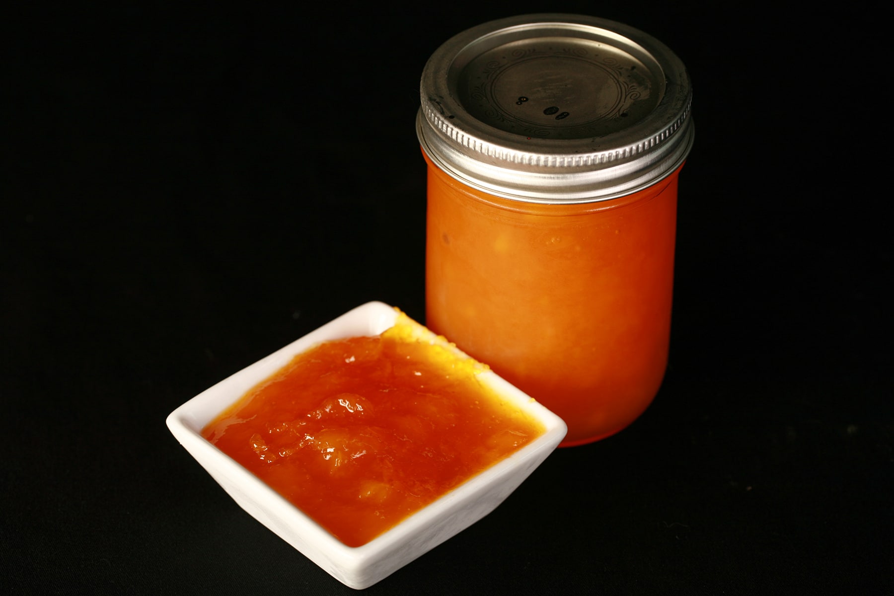 A jar of small batch mango peach jam, next to a small white bowl of the jam.