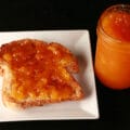 An open jar of mango peach jam next to a slice of toast on a plate. The toast is generously spread with the jam.