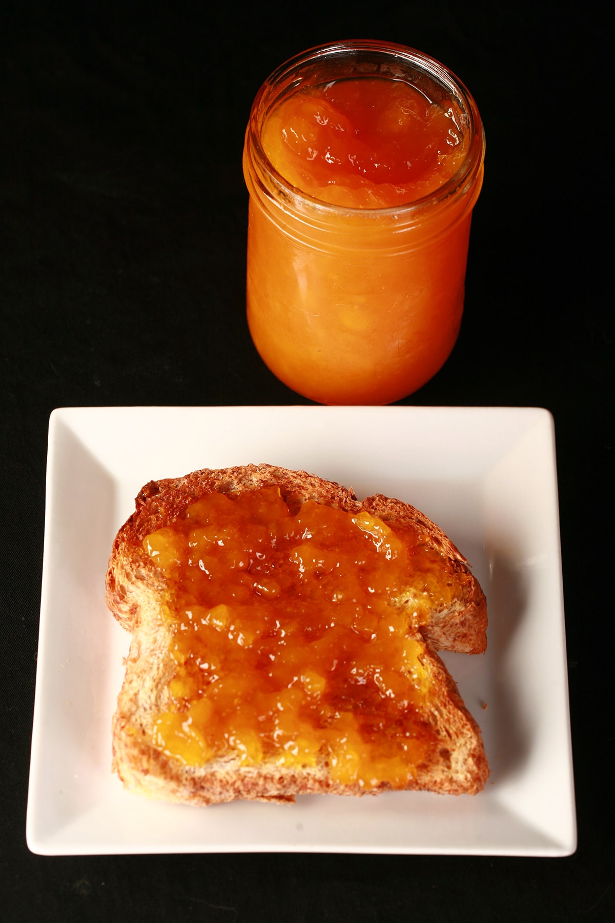 An open jar of mango peach jam next to a slice of toast on a plate. The toast is generously spread with the jam.