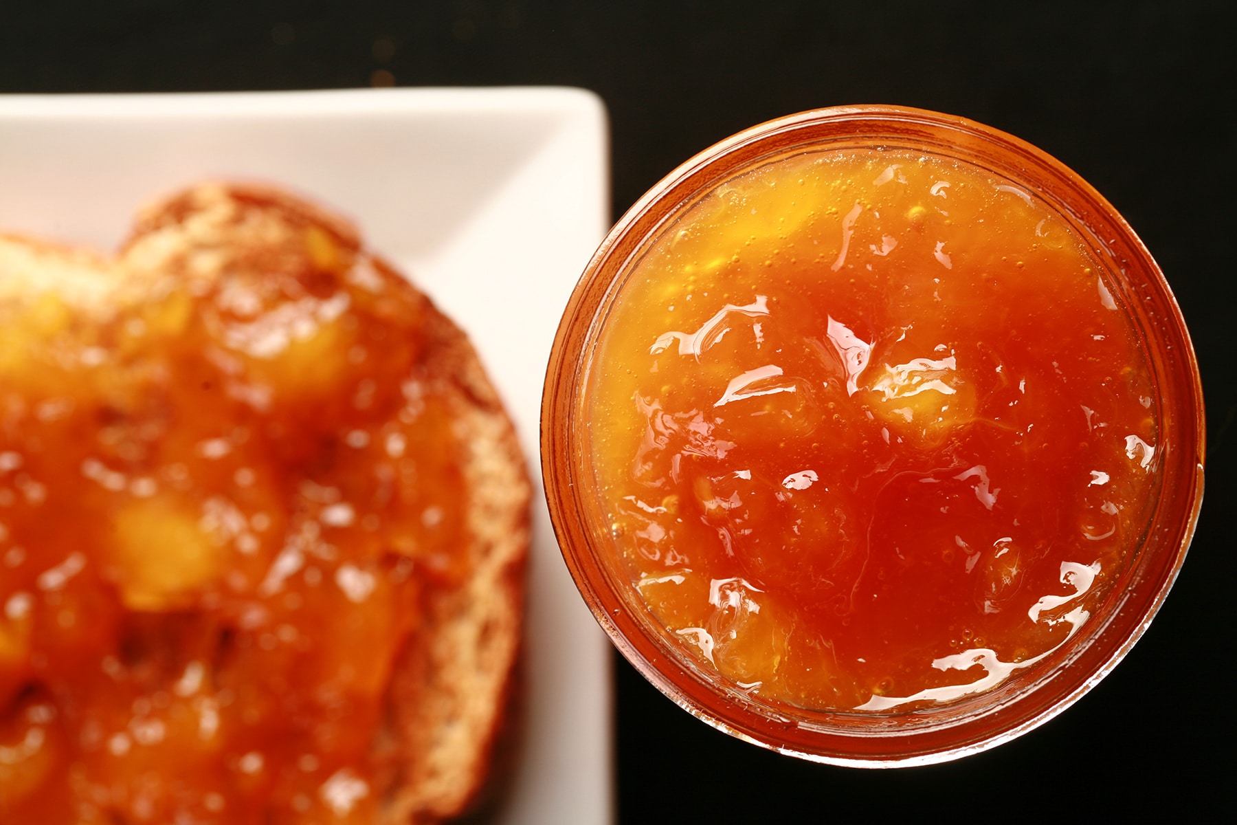 Close up photo of a small jar of orange coloured jam next to a piece of toast