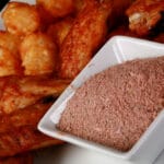 A small, square white bowl of a deep orange-red dry rub, on a larger plate of wings and tater tots. The wings are coated in the same red dry rub.