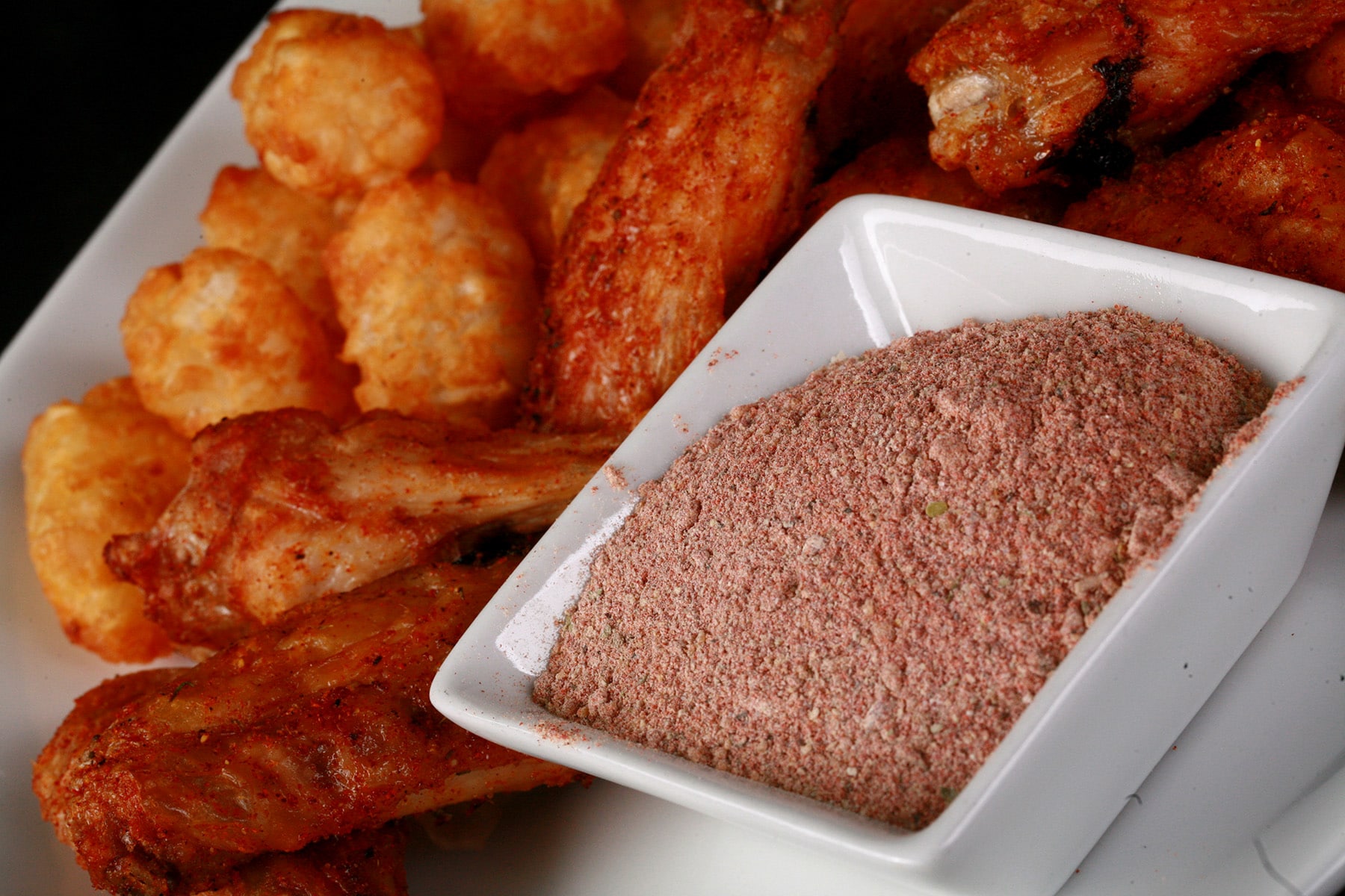 A small, square white bowl of a deep orange-red dry rub, on a larger plate of wings and tater tots. The wings are coated in the same red dry rub.