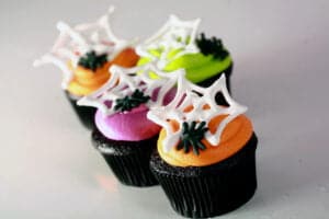 4 black velvet Halloween cupcakes frosted with brightly coloured icing - lime green, electric purple, and orange - are shown topped with royal icing spider webs and spiders.