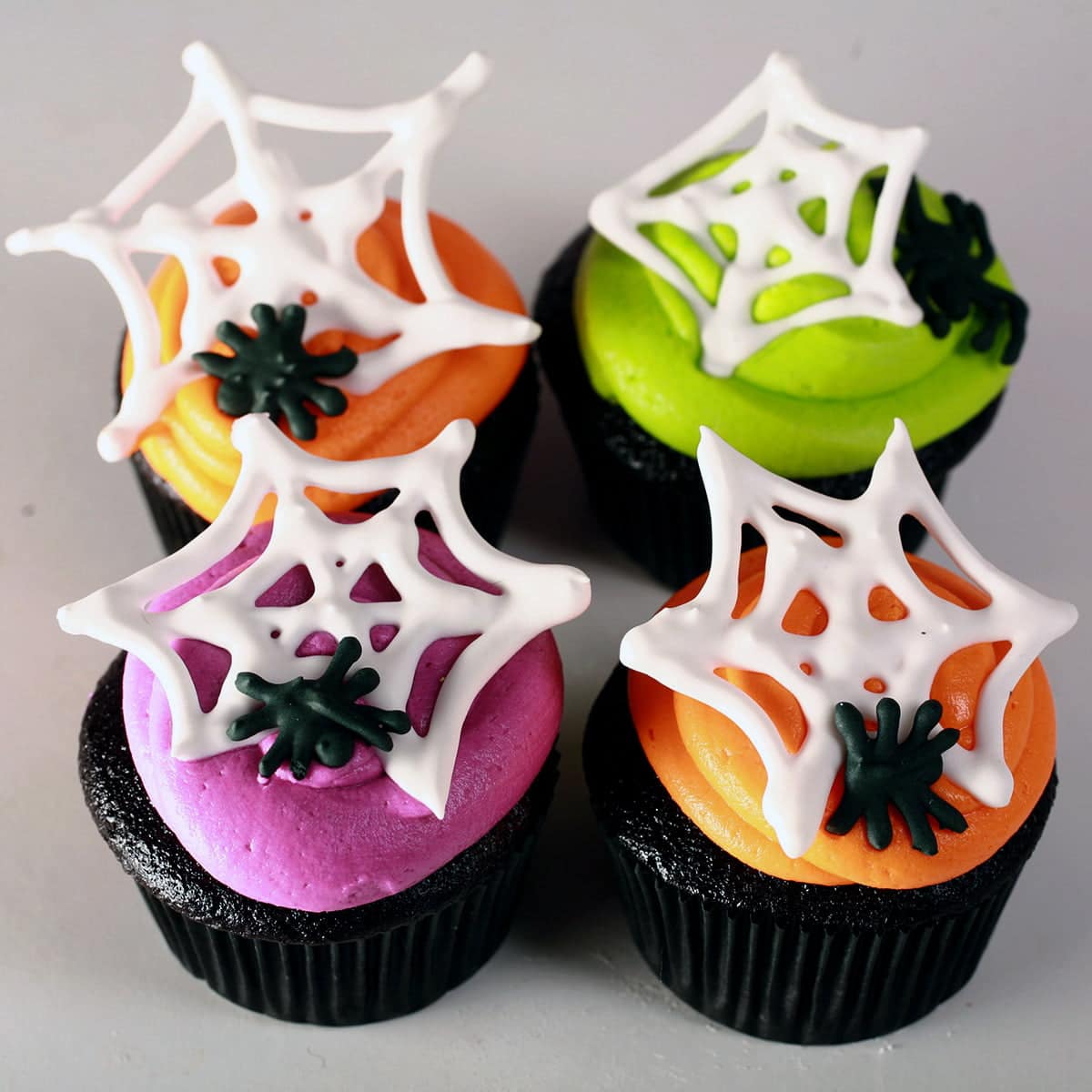 4 black velvet cupcakes frosted with brightly coloured icing - lime green, electric purple, and orange - are shown topped with royal icing spider webs and spiders.