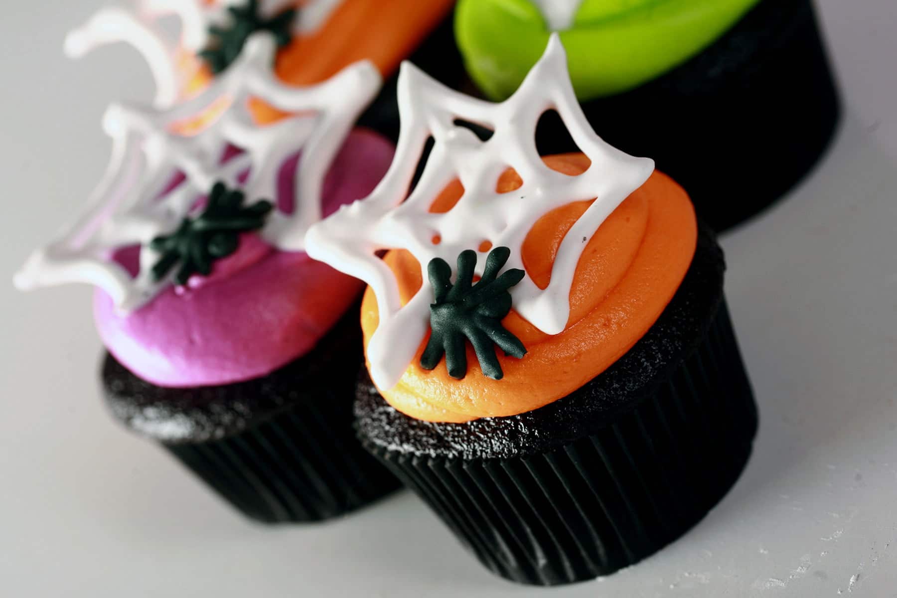 4 black velvet cupcakes frosted with brightly coloured icing - lime green, electric purple, and orange - are shown topped with royal icing spider webs and spiders.
