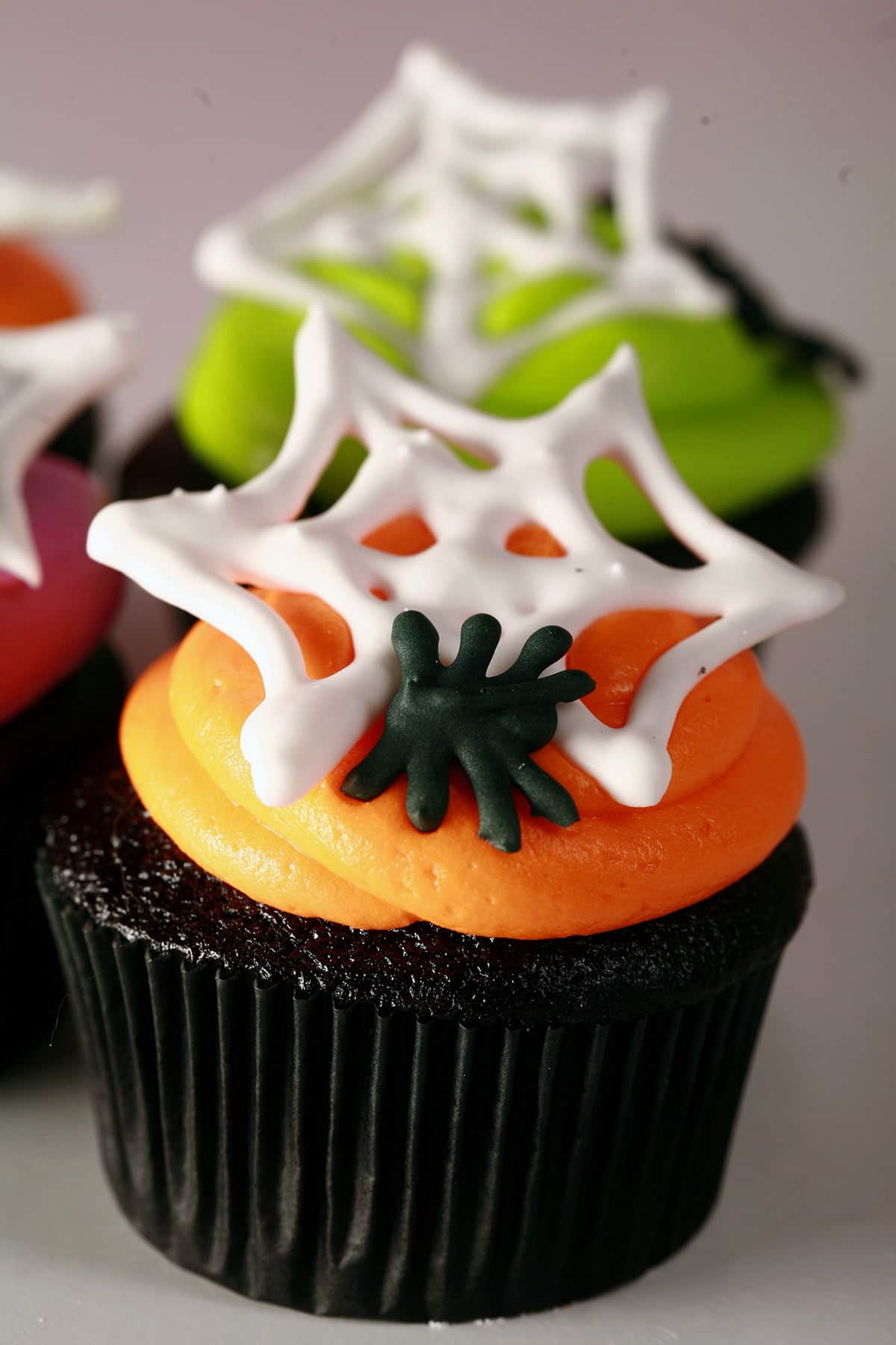 4 black velvet Halloween cupcakes frosted with brightly coloured icing - lime green, electric purple, and orange - are shown topped with royal icing spider webs and spiders.