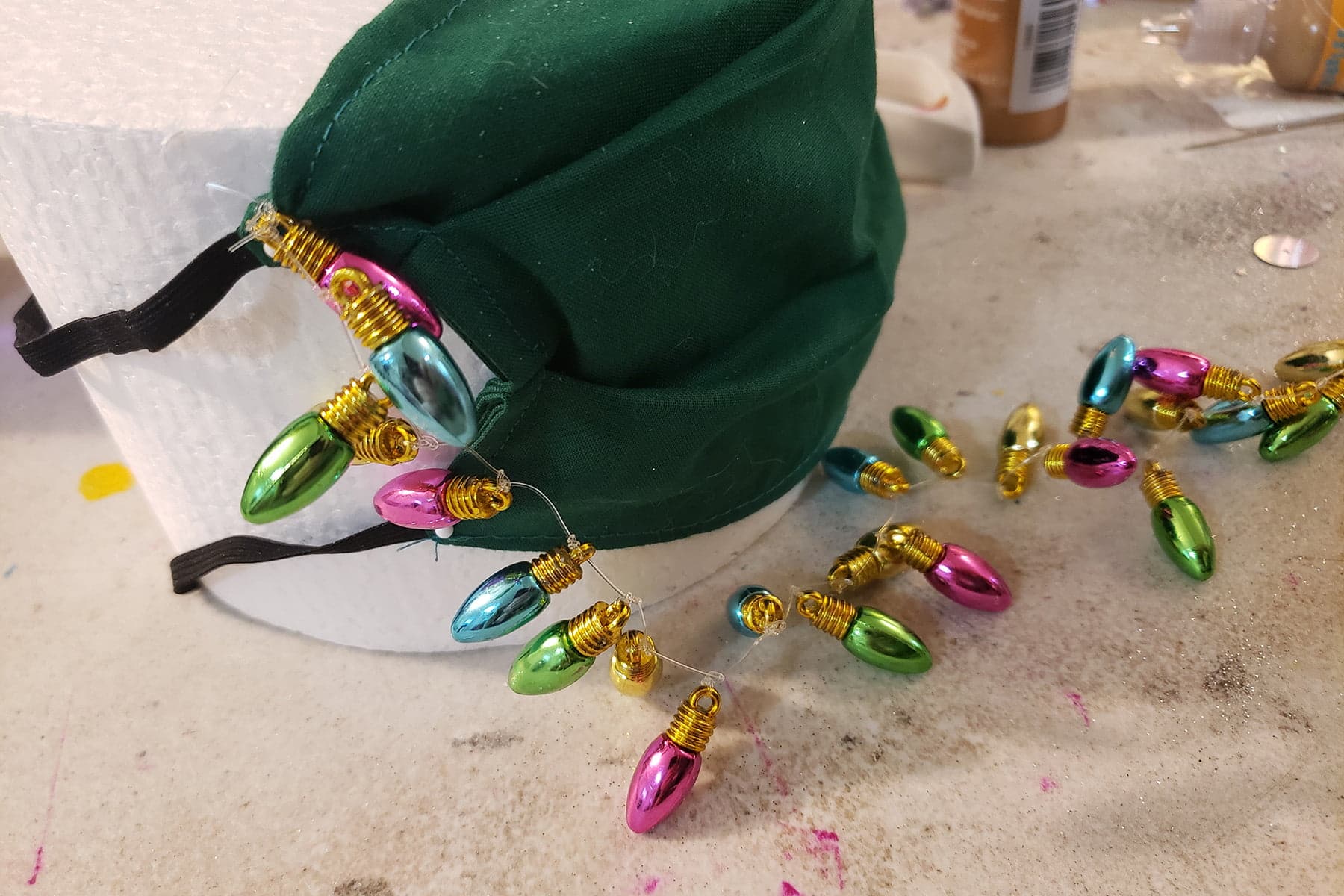 A green mask attached to a round of styrofoam.  A string of beads that look like Christmas tree lights is secured to one corner, and laying on the table in front of the mask.
