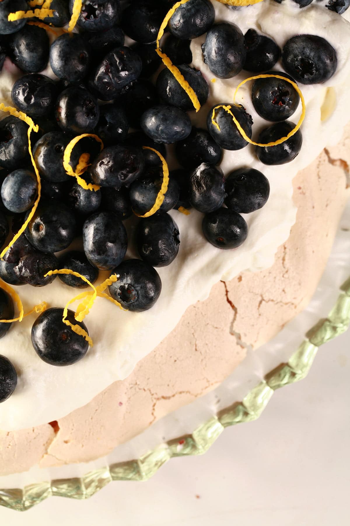 Close up view of a blueberry pavlova. The meringue is light purple, it is topped with whipped cream, blueberries, and curls of lemon zest.