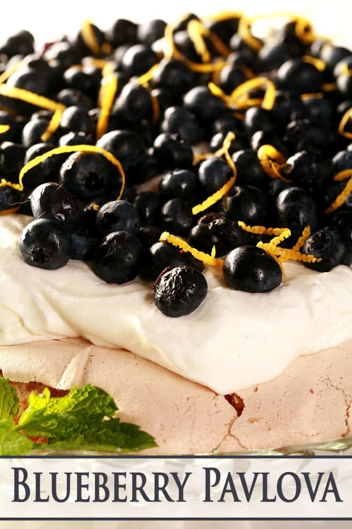 Close up view of a blueberry pavlova. The meringue is light purple, it is topped with whipped cream, blueberries, and curls of lemon zest.