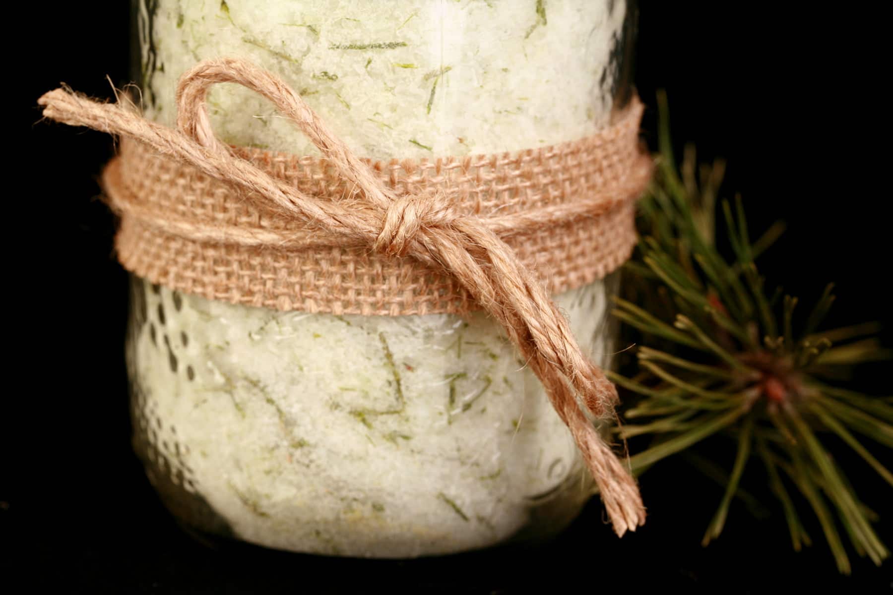 A mason jar filled with a pale green salt that has green flecks of pine needles throughout. It is wrapped with a burlap ribbon and tied with twine.