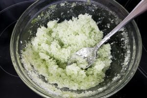 A glass bowl with pale green salt in it. There are flecks of pine needles throughout the salt.