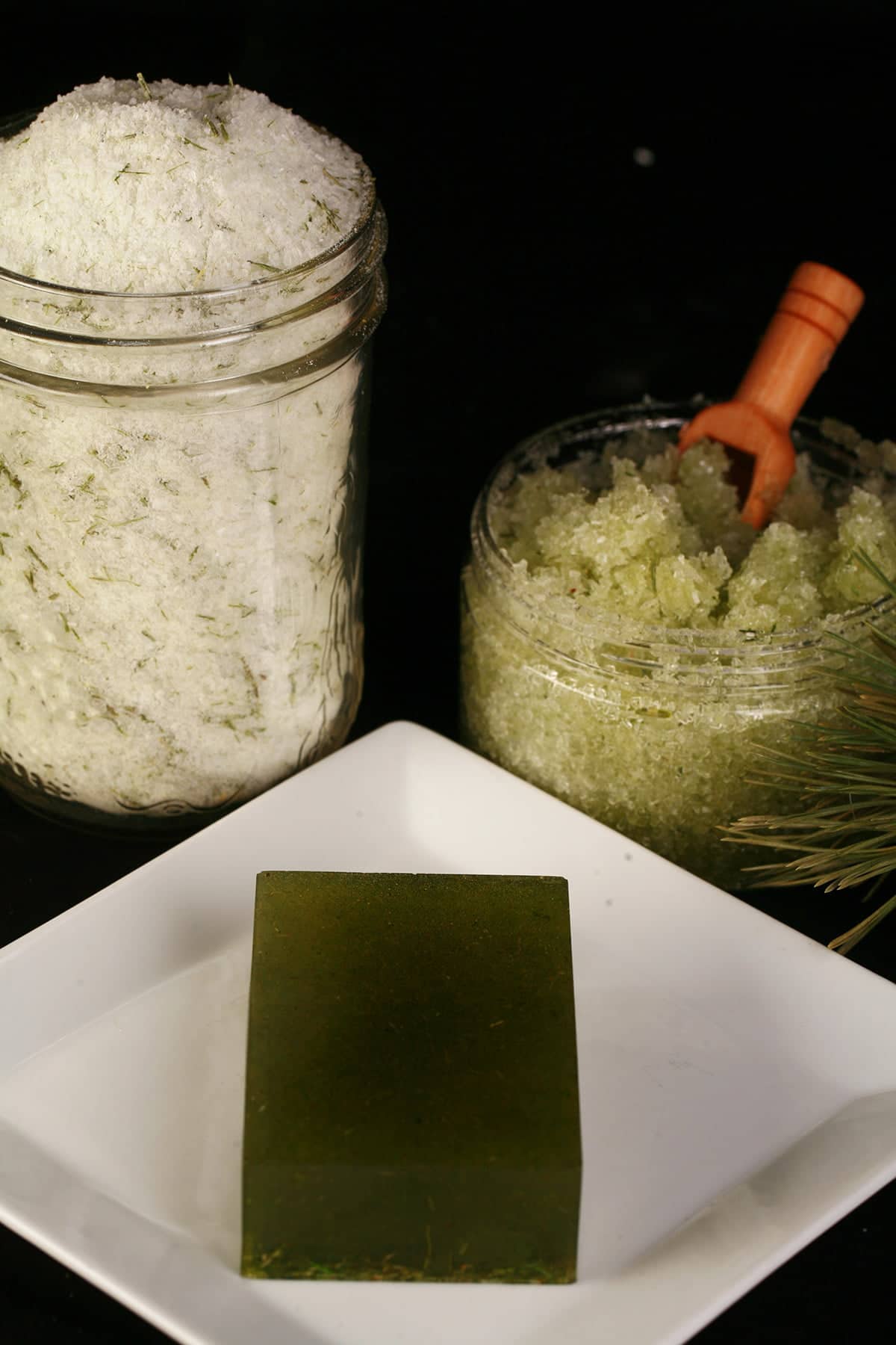 A trio of cedar bath products - a mason jar with bath salts, a smaller jar with green salt scrub, and a green bar of soap. All have flecks of preen pine needles in them.