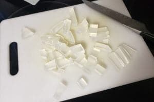 A cutting board with a cubes of clear soap piled on it. There is a large knife in the background,