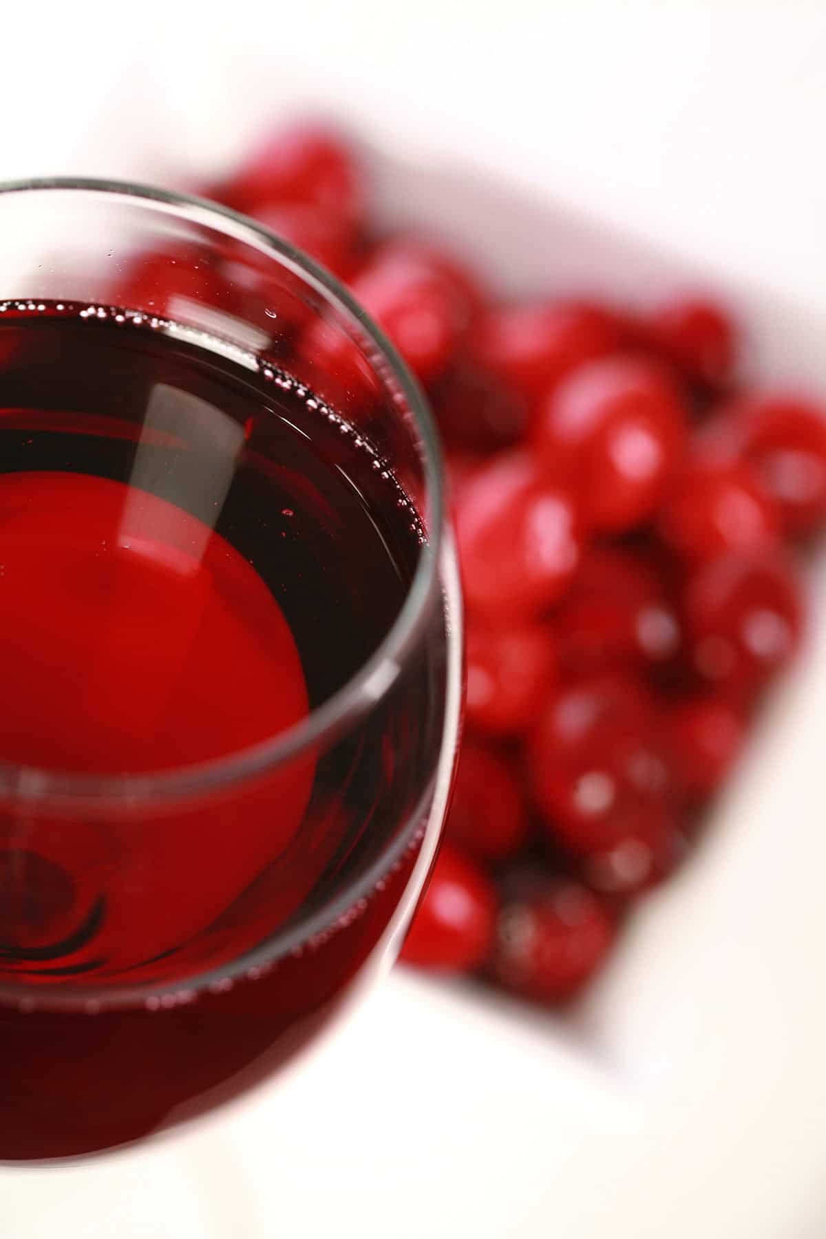 A glass of red wine -made with this cranberry wine recipe - is pictured next to a small bowl of fresh cranberries, against a white background.