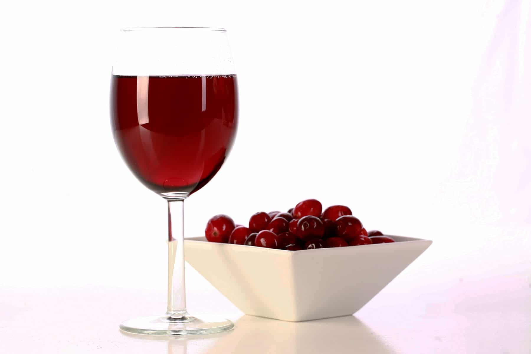 A glass of red wine is pictured next to a small bowl of fresh cranberries, against a white background.