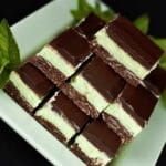 A plate of Creme de Menthe Nanaimo Bars - a 3 layered bar. The top and bottom layers are chocolate, and the middle layer is a green buttercream. They are on a green plate, garnished with a sprig of fresh mint.