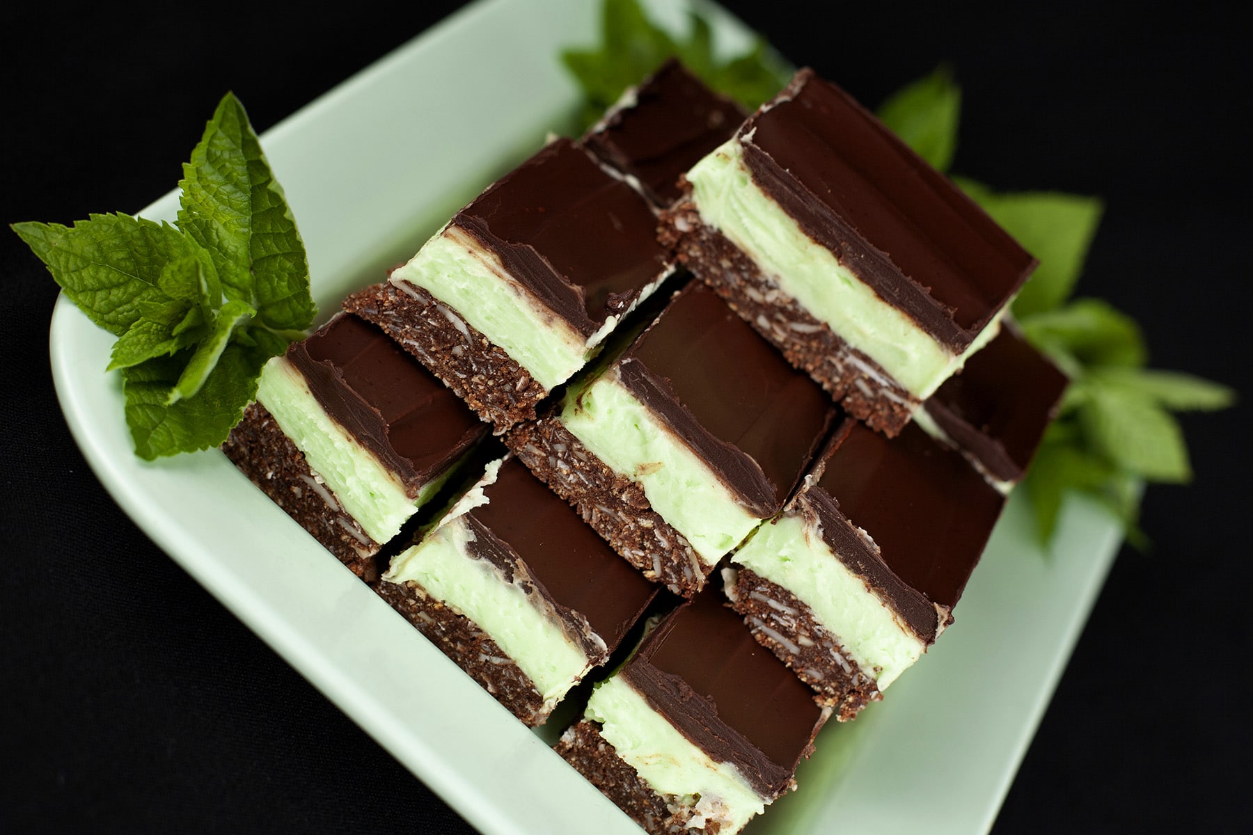 A plate of Creme de Menthe Nanaimo Bars - a 3 layered bar. The top and bottom layers are chocolate, and the middle layer is a green buttercream. They are on a green plate, garnished with a sprig of fresh mint.