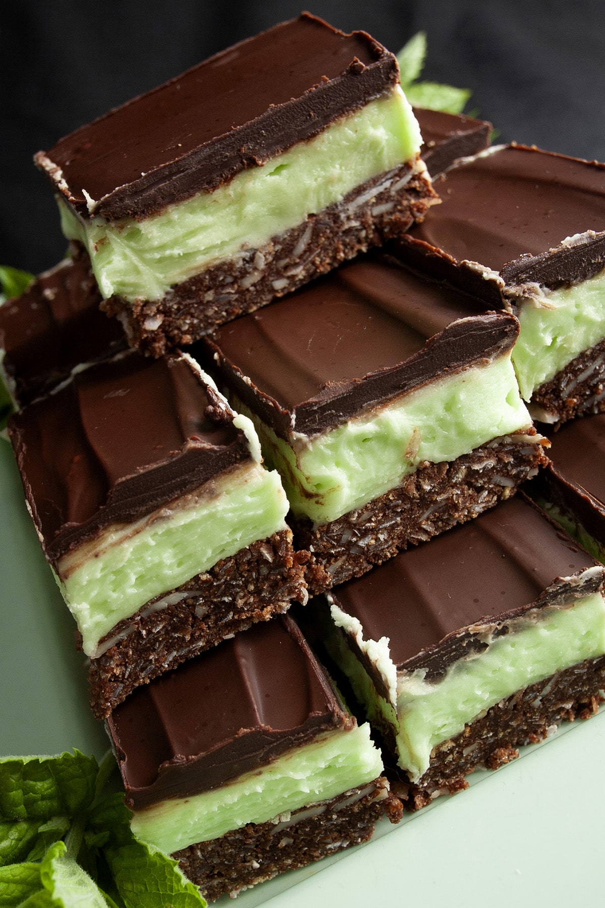 Close up view of a plate of Creme de Menthe Nanaimo Bars - a 3 layered bar. The top and bottom layers are chocolate, and the middle layer is a green buttercream. They are on a green plate, garnished with a sprig of fresh mint.