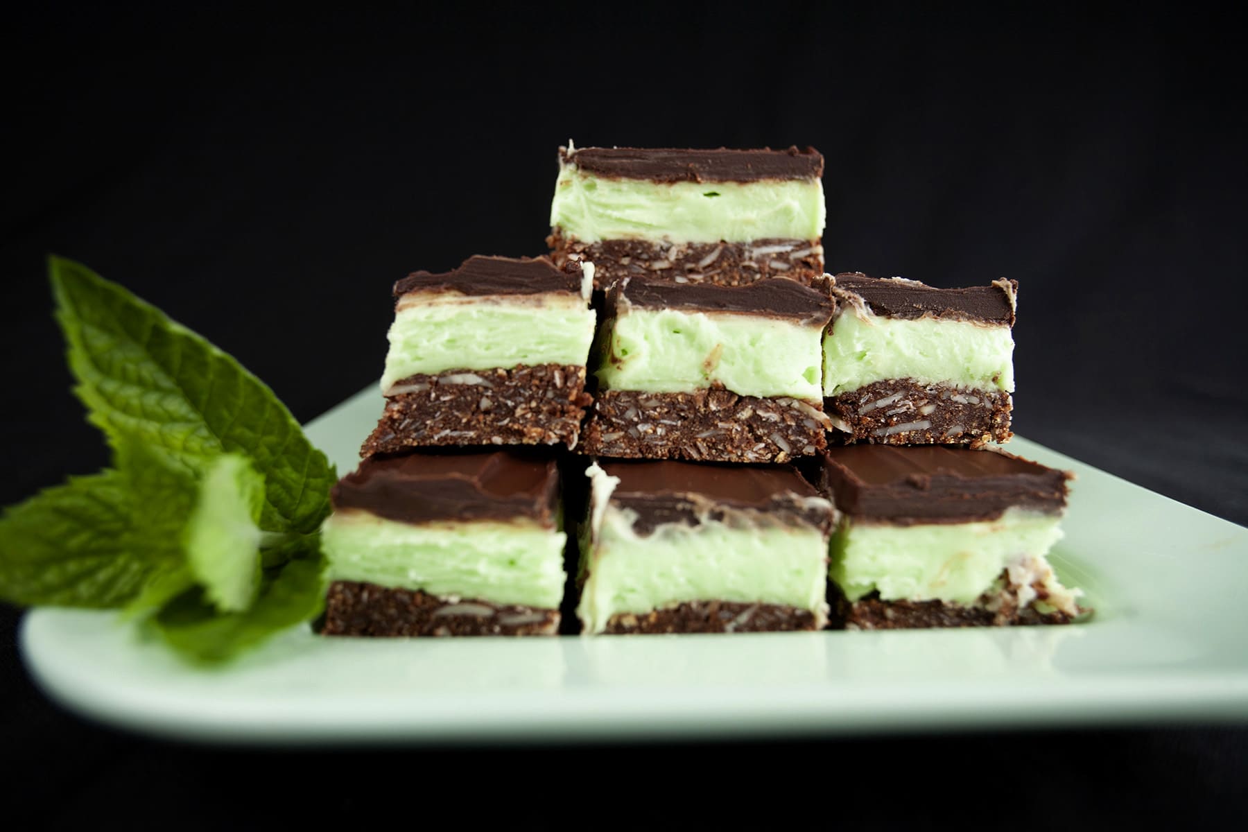 A plate of Creme de Menthe Nanaimo Bars - a 3 layered bar. The top and bottom layers are chocolate, and the middle layer is a green buttercream. They are on a green plate, garnished with a sprig of fresh mint.