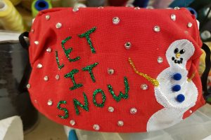 A red mask has a white snowman, and the words "Let it new" painted in green glitter. The whole mask is covered with clear rhinestones.