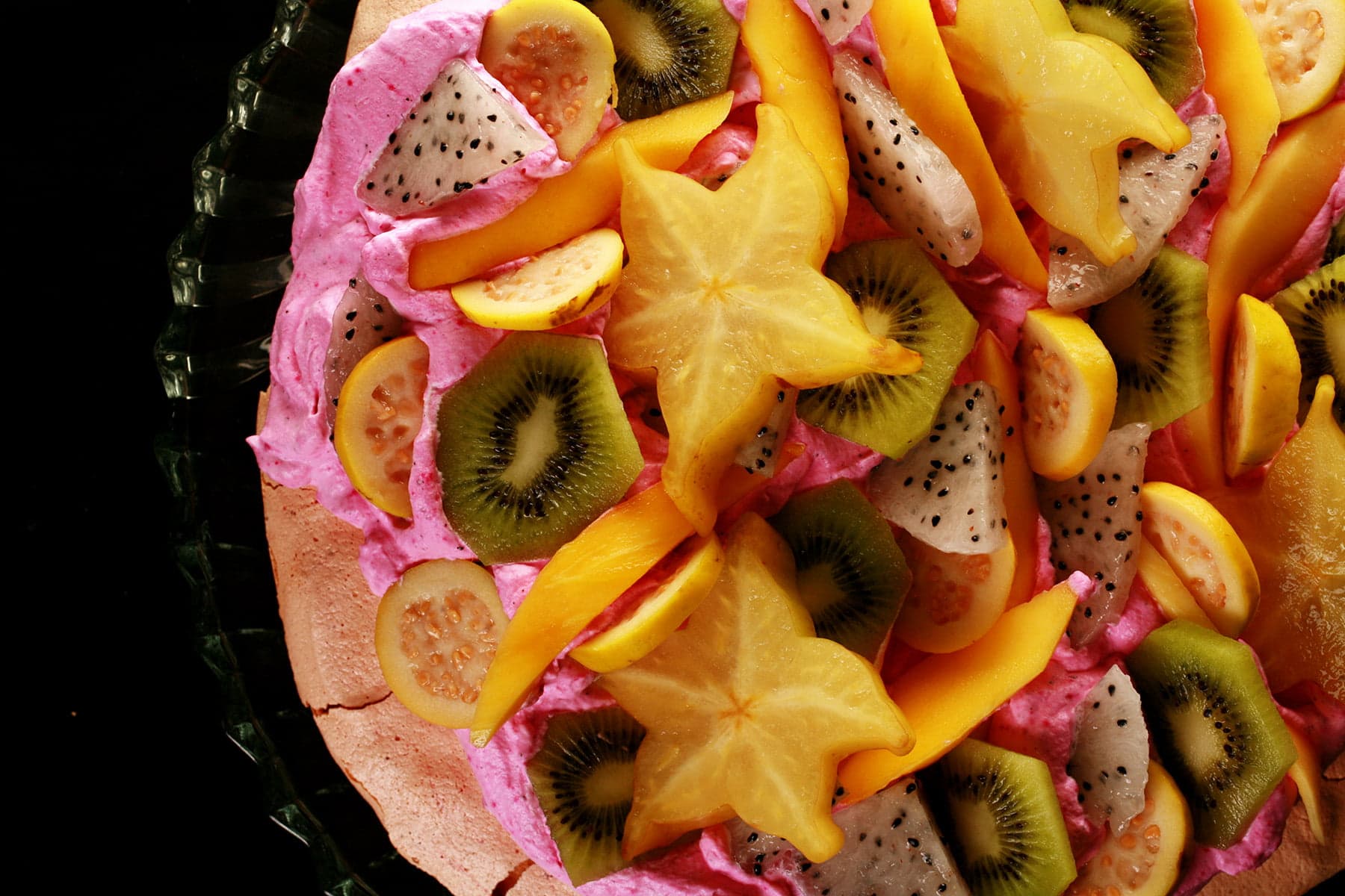 Close up view of a colourful pavlova. It has a light pink meringue base, bright pink whipped cream, and is topped with dragon fruit, star fruit, guava, and mango.