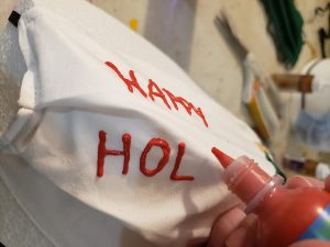 A white face mask is stretched over a round of styrofoam. A hand is piping the words "Happy Holidays" across the front, in red fabric paint.